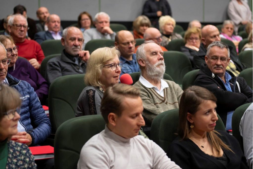 Kobieta siedząca wśród publiczności z mikrofonem w ręku, zadająca pytanie