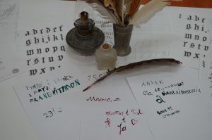 Na stole rozłożone są kartki z napisami zrobionymi gęsim piórem, lampa naftowa, szklany kałamarz i gęsie pióro