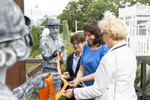 Trzy kobiety przecinają pomarańczową wstęgę, trzymaną przez dwie osoby przebrane za rzeźby.
