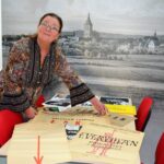Kobieta po lewej stronie stoi przy stole wyłożonym starymi plakatami i afiszami teatralnymi. Trzyma w rękach obrzeża jednego z plakatów. W tle nadruk z widokiem miasta Koszalin.