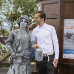 Elegancko ubrany mężczyzna z kobietą przebraną za rzeźbę na tle Archiwalnego Fotoplastikonu w Mielnie. Na dalszym planie drzewa i morze.