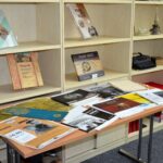 Stół wyłożony afiszami, publikacjami i dokumentami dotyczącymi działalności teatru "Dialog" w Koszalinie. W tle regał z zaprezentowanymi książkami.
