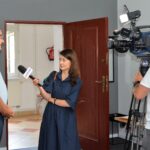 Trzy osoby w pokoju. Po lewej mężczyzna udzielający wywiadu, po środku dziennikarka z mikrofonem TV MAX, po prawej mężczyzna obsługujący kamerę.