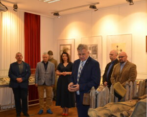 Sześć elegancko ubranych osób stoi w sali wystawowej. Na przodzie mężczyzna w garniturze łączy ręce i lekko pochyla głowę. Reszta stoi nieco z tyłu. Wszyscy mają poważne miny. W tle elementy wystawy - gabloty z eksponatami, manekin w stroju żołnierza, fotografie na ścianach i po prawej płotek z zawieszonymi elementami wyposażenia żołnierza.