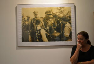 Duża fotografia żołnierzy i dziewcząt ubranych na ludowo wisi na ścianie w ramie. Po prawej stronie stoi kobieta zwrócona z półprofilu. Podpiera głowę ręką i patrzy w dal z delikatnym uśmiechem.
