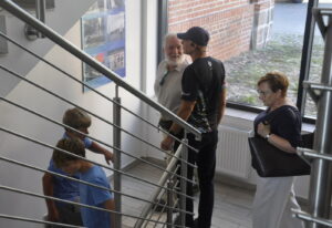 Klatka schodowa. Na półpiętrze dwóch mężczyzn przed zawieszoną na ścianie planszą ze starymi zdjęciami na niebieskim tle. Po prawej stronie kobieta z dużą czarną torebką. Na schodach poniżej półpiętra dwóch chłopców w niebieskich koszulkach widzianych przez metalowe poziome pręty poręczy. W tle duże okno z widocznym dziedzińcem.