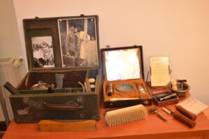 Na stoliku z pomarańczowym nakryciem leżą eksponaty. Są to elementy wyposażenia żołnierza. Po lewej otwarty kuferek z przyczepionymi po wewnętrznej stronie wieczka zdjęciami. Po prawej przybory toaletowe: szczoteczka, przybory do golenia, lusterko.