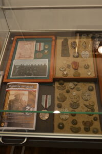 Fragment ekspozycji. W gablocie ułożone stare pamiątki wojenne: zdjęcia, medale i resztki nieśmiertelników.