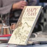 Książka Mapy i plany Koszalina do 1945 roku stoi na stojaku na stole. W ten sposób została wyeksponowana.