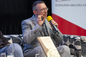 Mężczyzna z żółtym mikrofonem w ręce siedzi i opowiada.