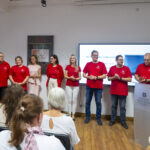 Kilka osób w czerwonych koszulkach stoi w sali przed widownią. Po prawej mównica z napisem Archiwa Państwowe Archiwum Państwowe w Koszalinie. Za mównicą kobieta w sukience. Wszyscy się uśmiechają.
