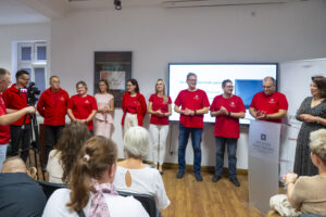 Kilka osób w czerwonych koszulkach stoi w sali przed widownią. Po prawej mównica z napisem Archiwa Państwowe Archiwum Państwowe w Koszalinie. Za mównicą kobieta w sukience. Wszyscy się uśmiechają.