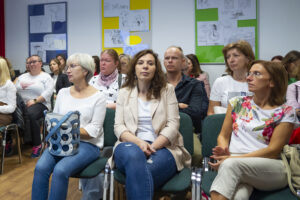 Spora grupa osób siedzi na krzesłach w pomieszczeniu i słucha wykładu. Na ścianach trzy kolorowe plansze z rysunkami satyrycznymi.