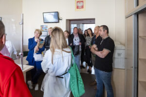 Kilkanaście osób stoi w małym pomieszczeniu i słucha. Kobieta w niebieskiej garsonce i blond włosach zadaje pytanie.