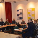 Kilka osób siedzi przy stołach ustawionych w podkowę. Słuchają wystąpienia.