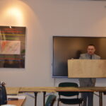 Mężczyzna w garniturze stoi za stołem i trzyma w rękach starą, rozłożoną mapę. Opowiada.