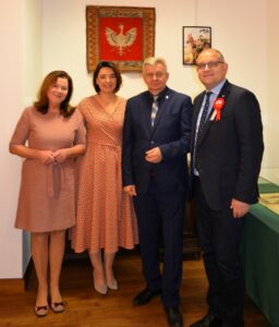 Cztery elegancko ubrane osoby - dwie kobiety i dwóch mężczyzn - stoją i pozują do zdjęcia na tle materiału z orłem.