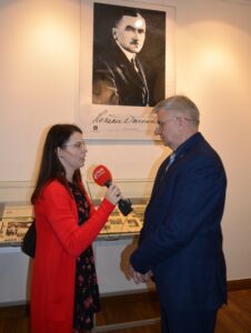 Kobieta w czerwonym swetrze z mikrofonem w ręku stoi na przeciwko mężczyzny w garniturze. W tle wisi na ścianie portret Romana Dmowskiego.