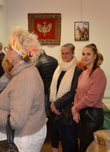 Kilka osób w sali wystawowej. Z przodu kobieta w blond włosach i przepasce widziana od tyłu. Na drugim planie dwie kolejne kobiety delikatnie uśmiechnięte stoją ze skrzyżowanymi rękoma.