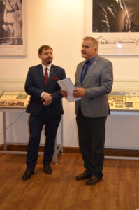 Dwóch elegancko ubranych mężczyzn stoi na tle gablot. Jeden z nich trzyma kartki papieru i opowiada.