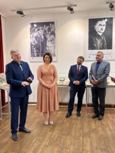 Cztery osoby na środku sali wystawowej. Stoją i uśmiechają się. W tle gabloty z eksponatami, a na ścianie czarno-białe zdjęcia Ojców Niepodległości.