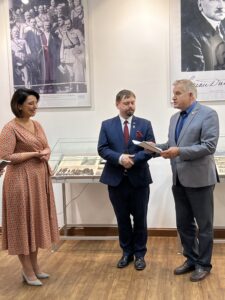Trzy osoby stoją na środku sali wystawowej. Po lewej kobieta w sukience uśmiecha się. Po prawej dwóch mężczyzn w garniturach. Jeden z nich opowiada. W tle gabloty i zdjęcia na ścianach.