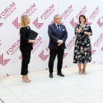 Trzy elegancko ubrane osoby stoją na tle ścianki z napisem Forum Koszalin. Kobieta po prawej stronie trzyma mikrofon.