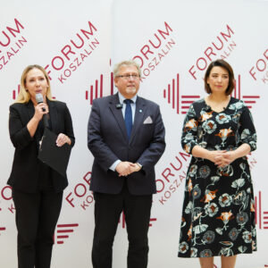 Trzy elegancko ubrane osoby stoją przed ścianką z napisem Forum. Kobieta po lewej stronie trzyma mikrofon.