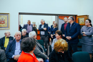 Grupa ludzi w pomieszczeniu czytelni. Większość elegancko ubrana. Część stoi, część siedzi. Mężczyzna w centralnej części zdjęcia śmieje się.