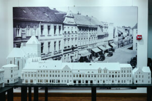 Biała makieta budynków z lat 30. XX wieku na tle starej fotografii ściennej ukazującej te same obiekty.