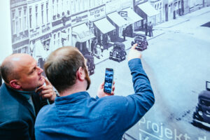 Dwóch mężczyzn stoi przed fotografią starych budynków i samochodów. Jeden z nich trzyma w jednej ręce telefon ze zdjęciem samochodu, a drugą ręką pokazuje ten samochód na fotografii ściennej. Drugi, po lewej, przygląda się temu, przykładając palce do brody.