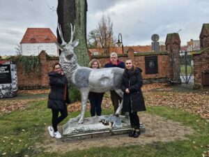 Czworo ludzi wokół srebrnej rzeźby Jelenia ustawionej na przed drzewem. Wokół trawa i jesienne liście. Z tyłu mur z cegieł.