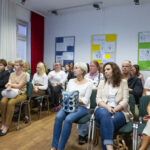 Kilkadziesiąt osób siedzi na krzesłach i słucha wykładu. Wokół na ścianach wiszą plansze z rysunkami satyrycznymi.