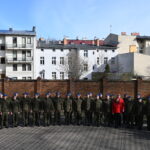 Grupa żołnierzy stoi w półkolu na dziedzińcu. Za nimi ceglany mur, a dalej budynki.