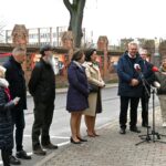 Dziewięcioro osób stoi na chodniku obok ulicy. Za nimi, po drugiej stronie ulicy, mur z czerwonej cegły, a na nim wystawa na dużych, podłużnych planszach.