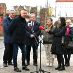 Siedem osób stoi na chodniku obok ulicy. Przed nimi mikrofony. Mężczyzna w ciemnym ubraniu i czarnych butach pochyla się nad jednym z rejestratorów dźwięku.