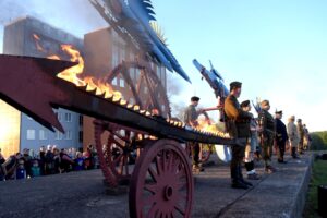 Płonące Ptaki Hasiora - rzeźba przedstawiająca mechaniczne ptaki na kołach. Przed rzeźbą ustawieni oficjele, za rzeźbą tłumek gapiów. W tle widać nowoczesny budynek składający się z dwóch prostokątnych bloków.